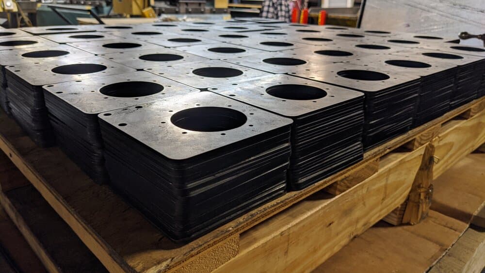 Stack of black metal plates with precision-cut circular holes, neatly arranged on a wooden pallet in an industrial setting.